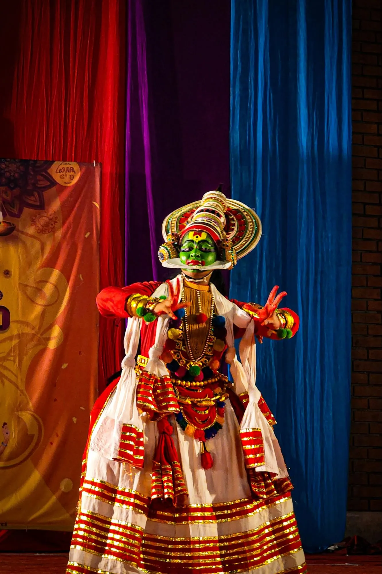 Attore di teatro-danza Kathakali con volto dipinto di verde e costume tradizionale rosso e bianco su sfondo di tendaggi colorati.
