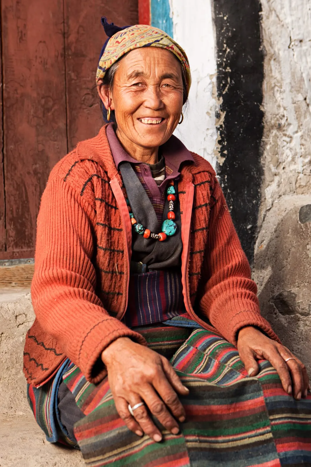 Una donna nepalese anziana sorridente seduta con abiti tradizionali, un cardigan arancione e gioielli turchesi, durante un trekking di gruppo in Nepal.