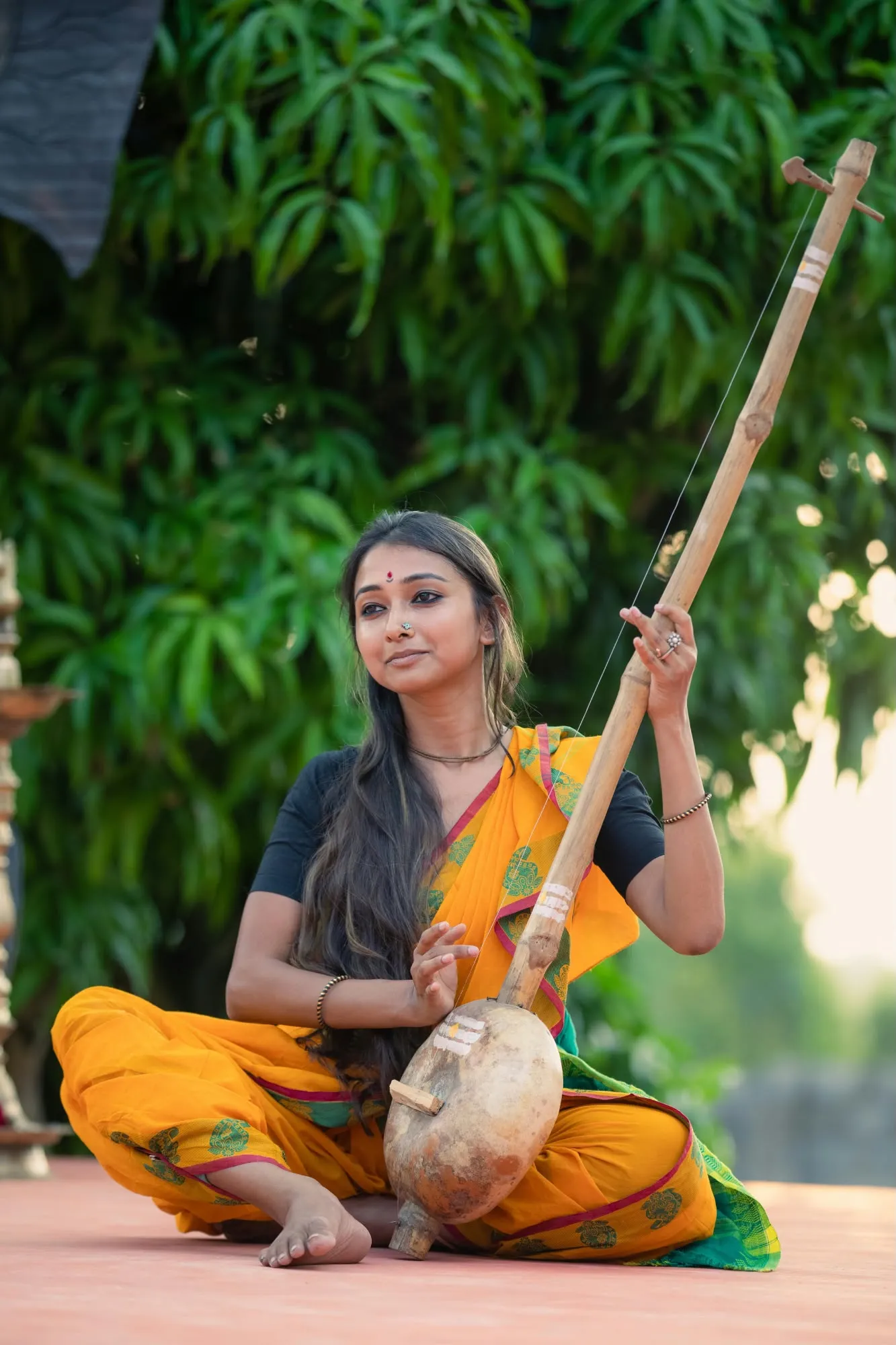 Una giovane donna indiana in sari giallo e nero seduta a terra mentre suona uno strumento musicale a corda fatto di zucca e bambù.