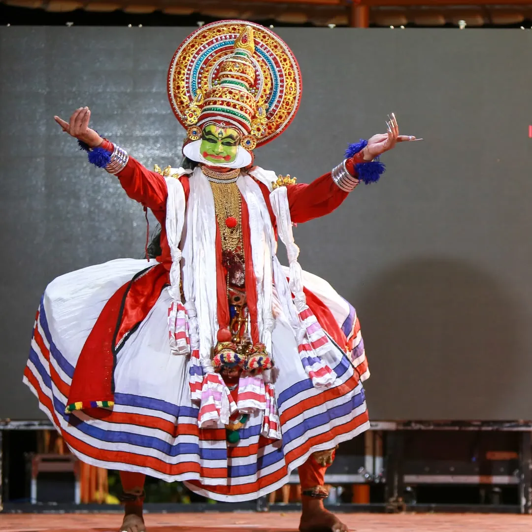 Ballerino di Kathakali sul palco con le braccia aperte, costume cerimoniale bianco e rosso e volto truccato di verde.