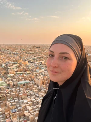 Primo piano di una donna con hijab nero che sorride davanti alla vista panoramica di Wadi Al-Salam a Najaf